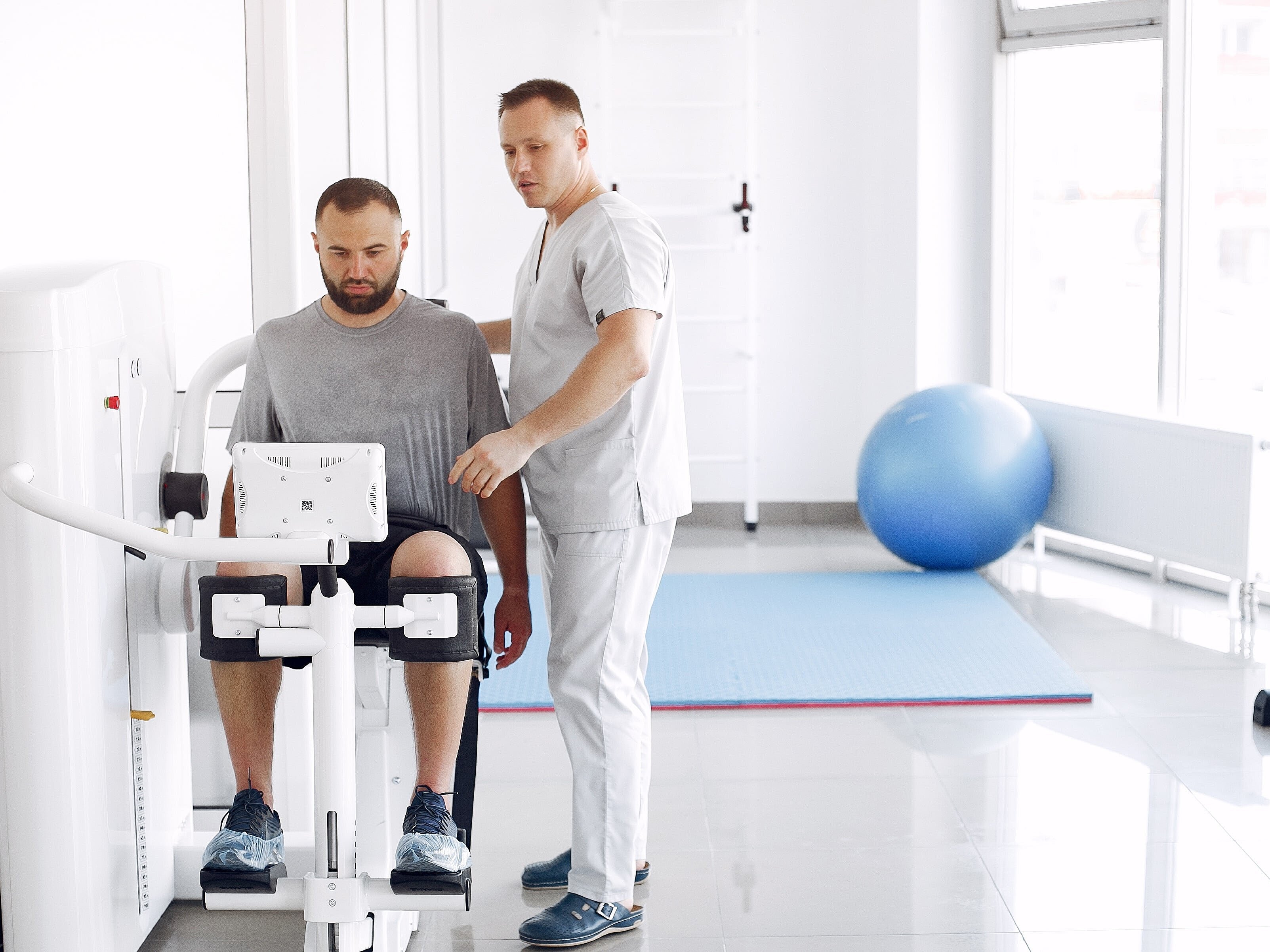 Ein Mann sitzt auf einem physiotherapeutischen Gerät. Daneben steht ein Mann, der ihm etwas erklärt.