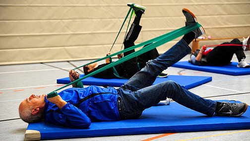 Menschen liegen auf Sportmatten und machen Übungen mit einem Gummiband.