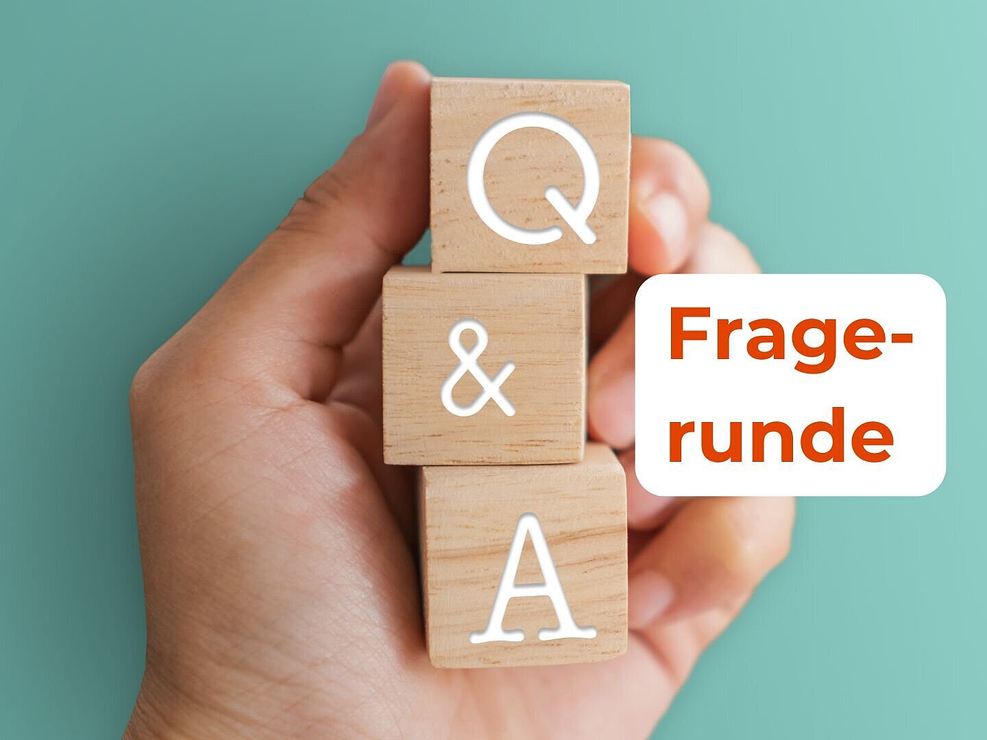 Eine Hand hält drei Holzwürfel, auf denen jeweils "Q", "&" und "A" steht. 