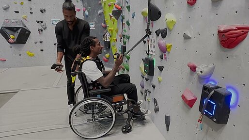 Kletterhalle Zwei Menschen, davon einer im Rollstuhl, vor einer Kletterwand.