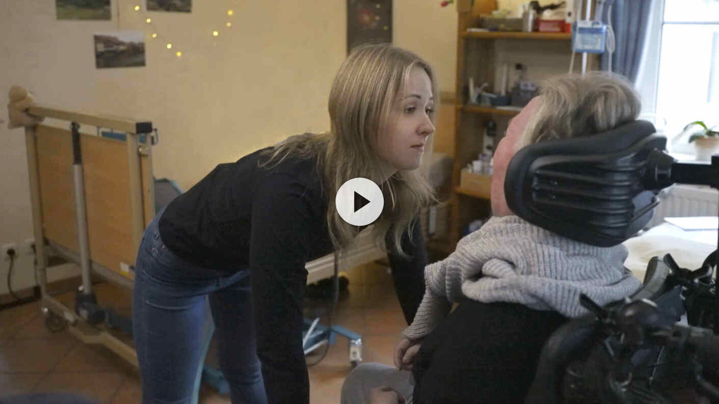 Eine junge Frau beugt sich zu einer im Rollstuhl sitzenden Frau hinunter.